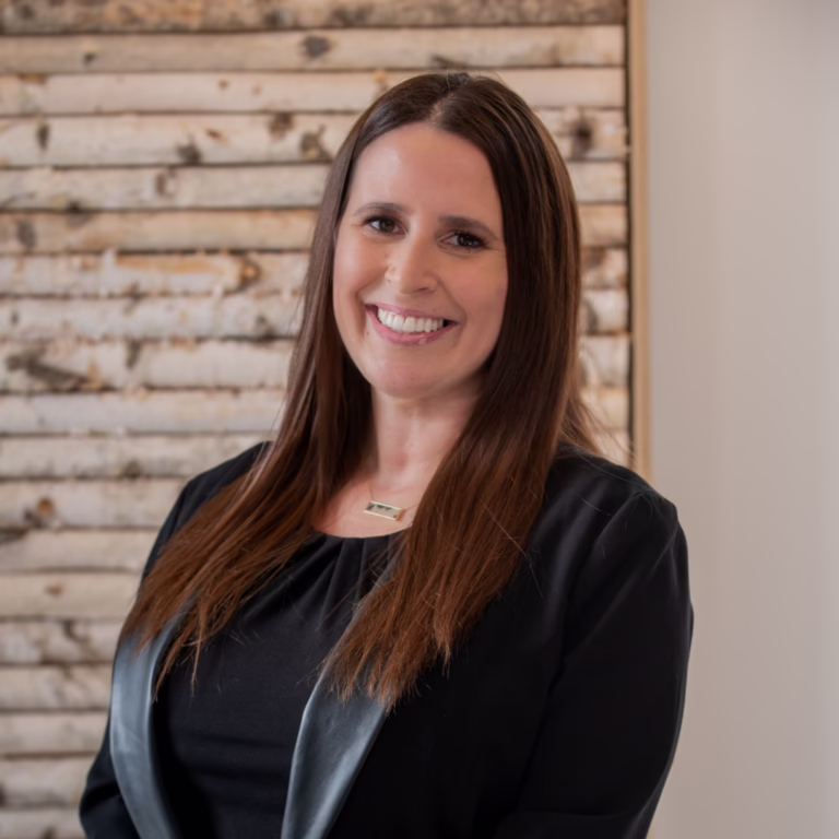 Smiling woman in a black blouse with leather trim, posing for a professional headshot in front of a wooden paneled wall.