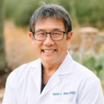 Smiling dentist in a white coat outdoors, with 'Randy L. Mow DDS' embroidered on the chest pocket area.