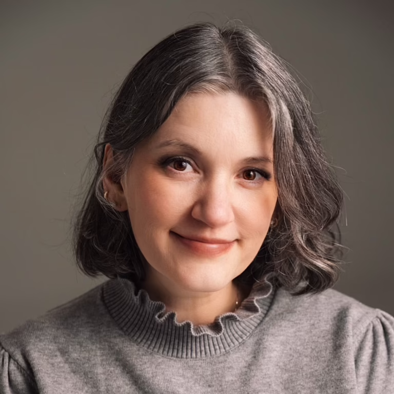 Smiling woman with short gray hair wearing a gray ribbed sweater against a neutral background.