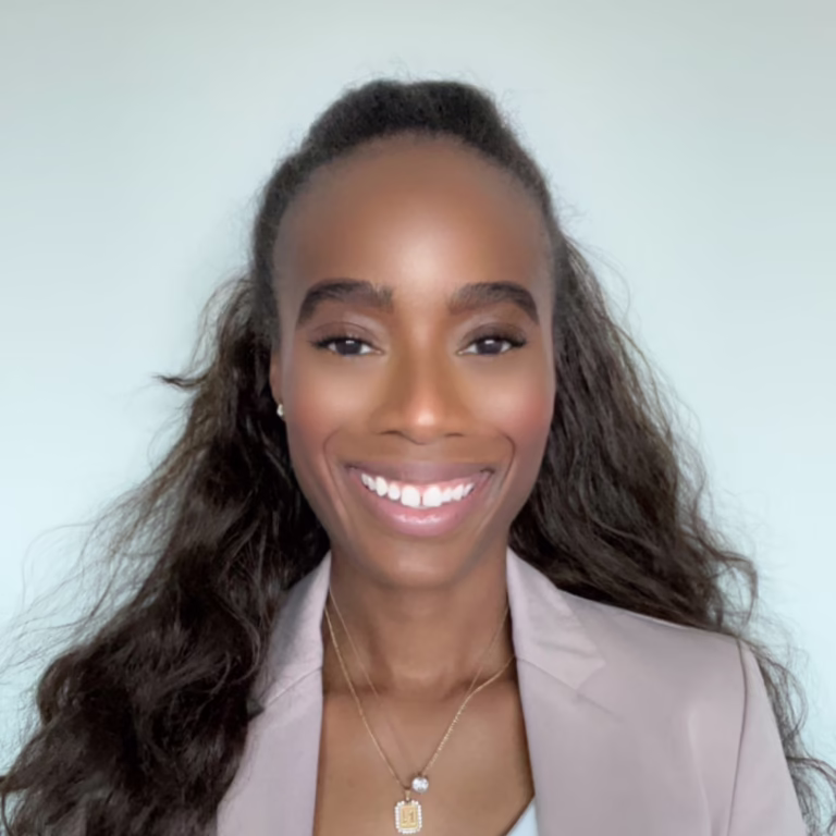 Portrait of a smiling Black woman with long wavy hair, wearing a light blazer and gold pendant necklace against a pale background.