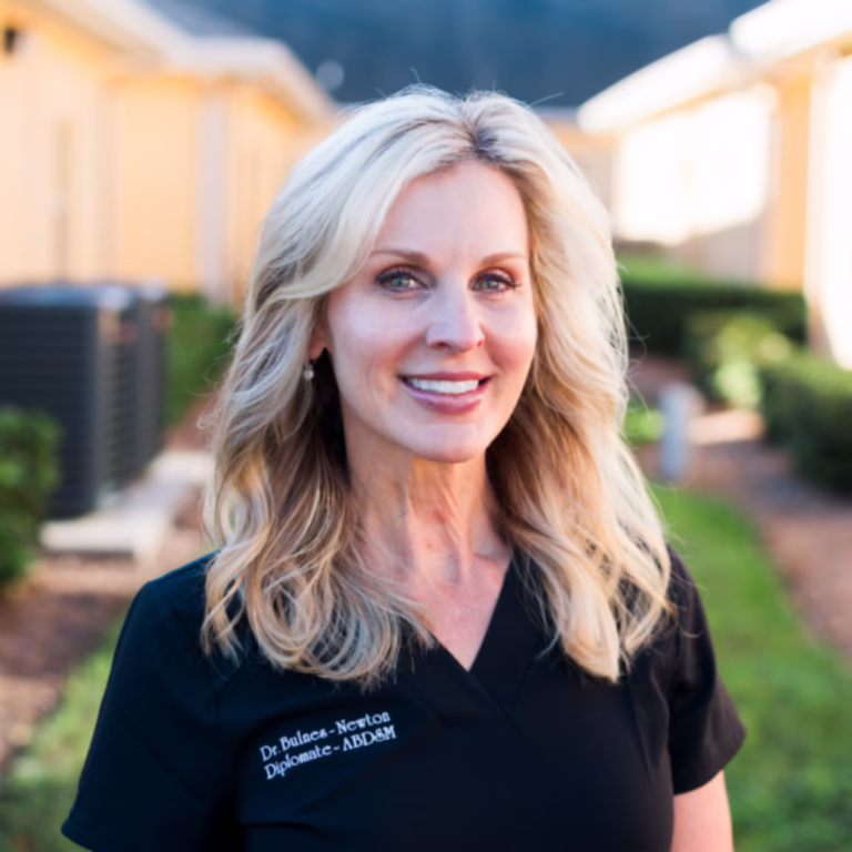 Smiling blonde woman in black scrubs standing outdoors in a sunny residential area.