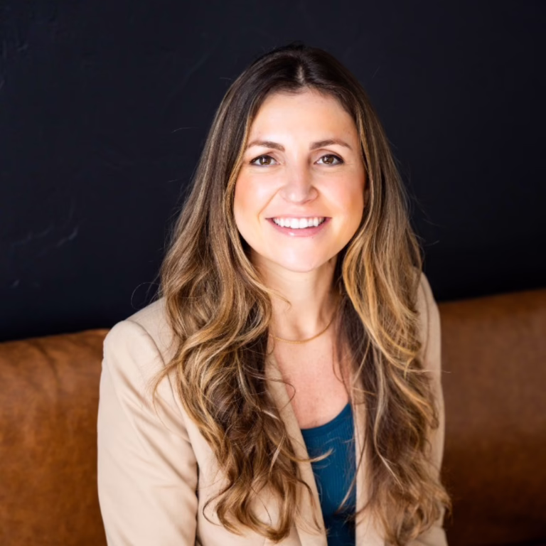 Smiling woman with long wavy hair in a beige blazer and teal top, seated on a brown couch.