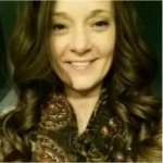 Close-up portrait of a smiling woman with wavy brown hair wearing a patterned paisley scarf in warm lighting.