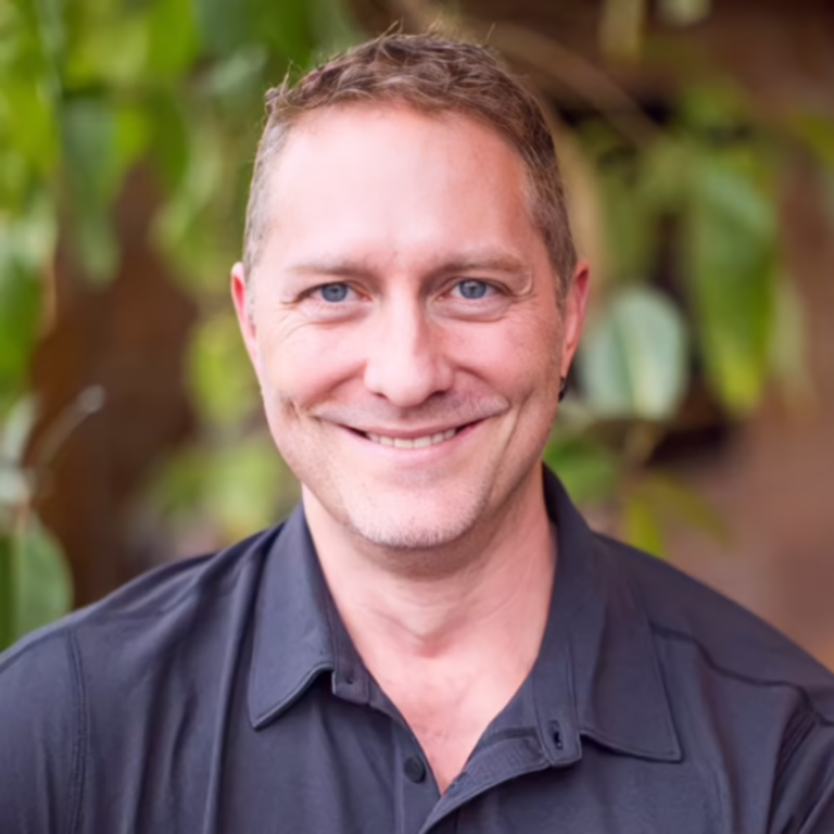 Smiling man with short light brown hair and blue eyes, wearing a dark shirt, outdoors with greenery in the background.