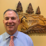 Smiling man in a blue checkered shirt and orange tie stands in front of a wooden carved sign that reads 'Okoniewski' with pine trees above it.