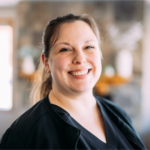 Smiling woman in black scrubs looking at the camera with a blurred indoor background, suggesting a healthcare setting.