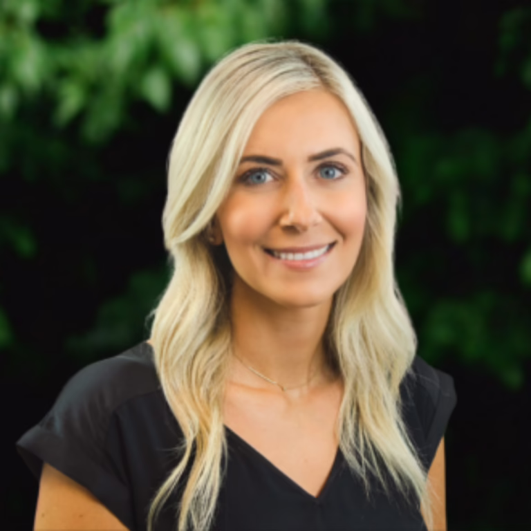 Portrait of a smiling blonde woman in a black top, with green foliage in the background.