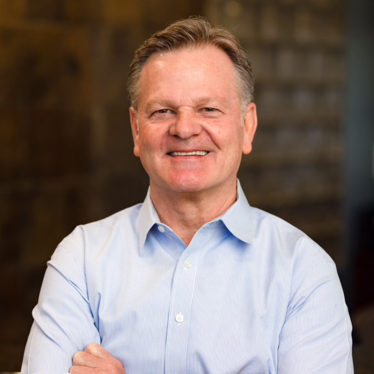 Professional headshot of a smiling man in a light blue button-down shirt with arms crossed, neutral office background