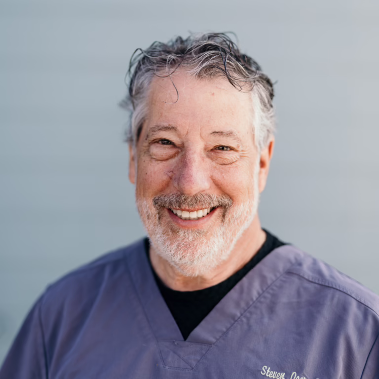 Smiling older man with gray hair and beard wearing purple medical scrubs outdoors, looking at the camera.