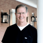 Smiling middle-aged male healthcare worker in black scrubs with glasses, standing in a brick-walled hallway.