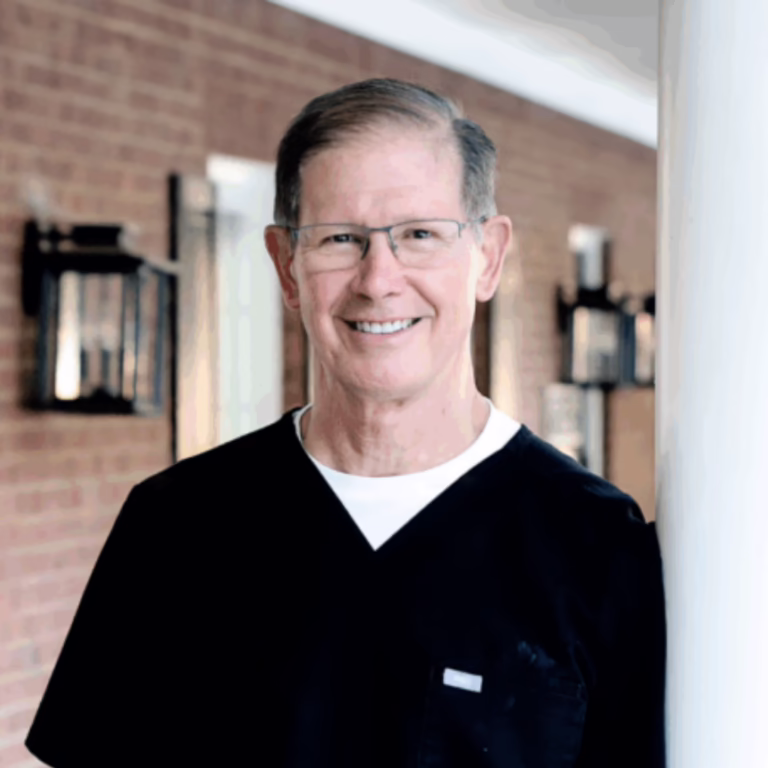 Smiling middle-aged male healthcare worker in black scrubs with glasses, standing in a brick-walled hallway.