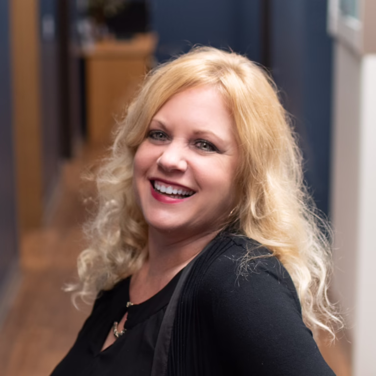 Smiling blonde woman in a black top standing in a hallway, looking at the camera.