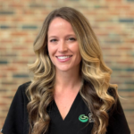 Smiling woman with long wavy blonde hair in a black scrub top, brick wall background, logo on chest.