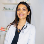 Smiling female healthcare professional in a white lab coat with a stethoscope around her neck in a clinical setting.