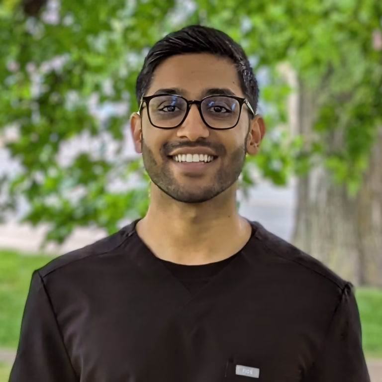 Smiling man with dark hair and glasses wearing black scrubs outdoors with green trees in the background, looking at the camera.