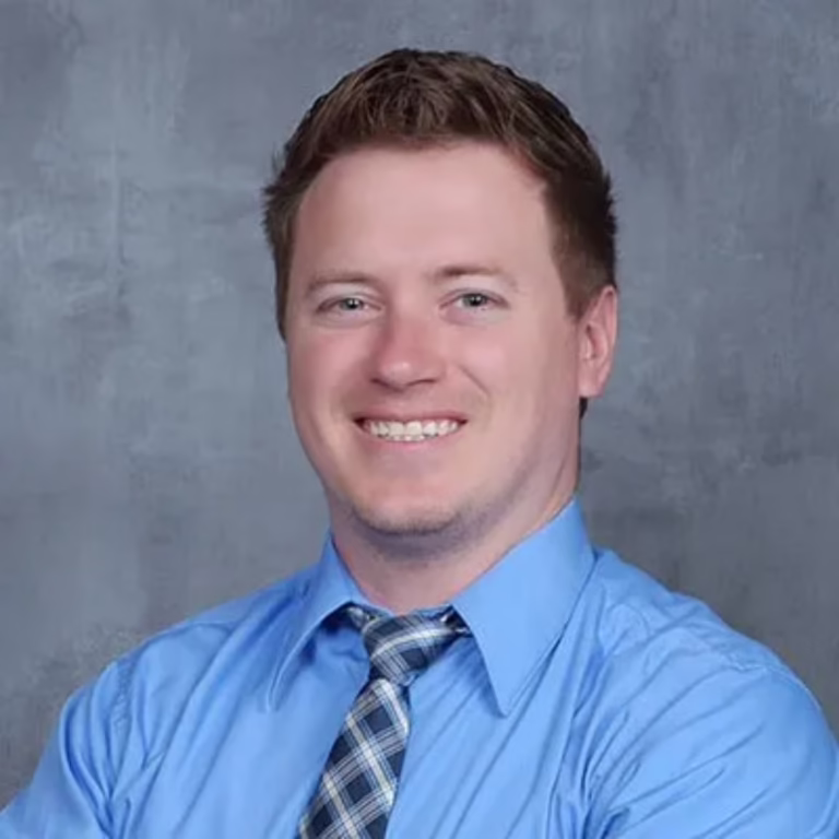 Friendly headshot of a man in a light blue shirt and plaid tie, smiling against a gray backdrop, professional portrait