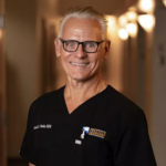 Smiling older man with gray hair and glasses in black scrubs, standing in a softly lit hallway.