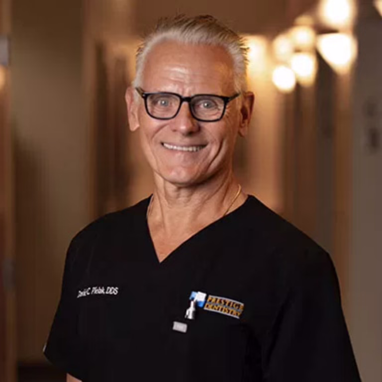 Smiling older man with gray hair and glasses in black scrubs, standing in a softly lit hallway.