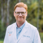 Portrait of a man with red hair and glasses wearing a white coat outdoors; the coat shows the name Aaron C. Polk D.D.S.
