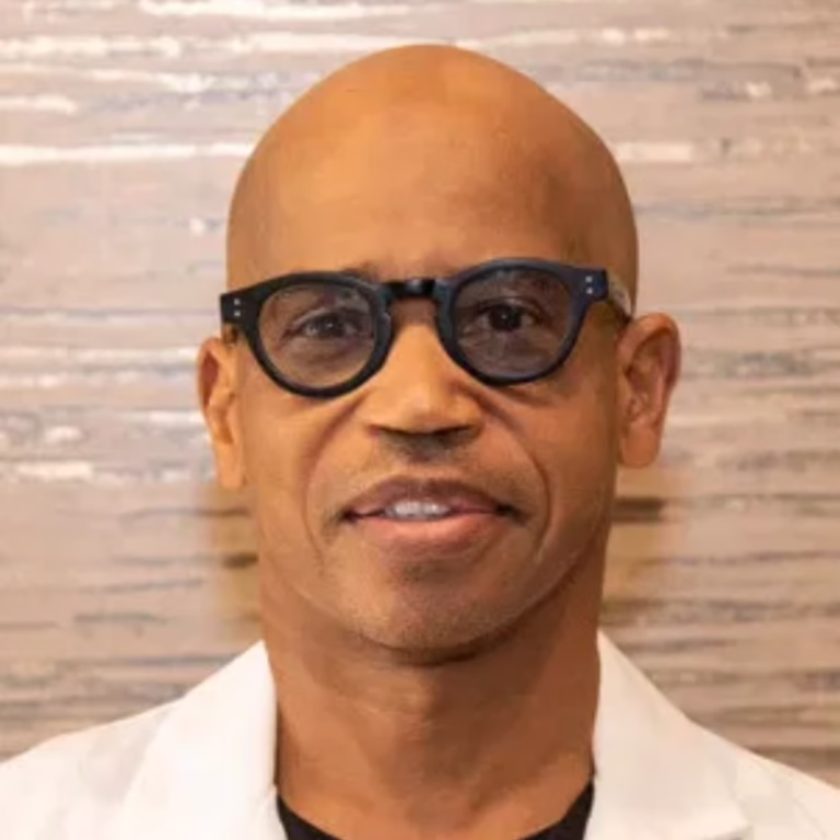 Smiling man wearing black-framed glasses and a white coat, standing against a light wooden background.