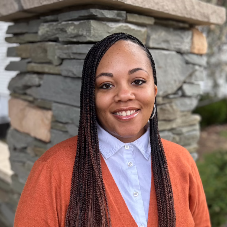 Smiling woman with long braided hair, wearing a light blue button-down shirt and orange cardigan, standing beside a stone wall outdoors.