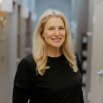 Smiling healthcare professional with long blonde hair wearing black scrubs in a clinical hallway.
