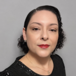 Portrait of a woman with short curly dark hair, red lipstick, wearing a black sequined top and a delicate gold necklace against a gray backdrop.