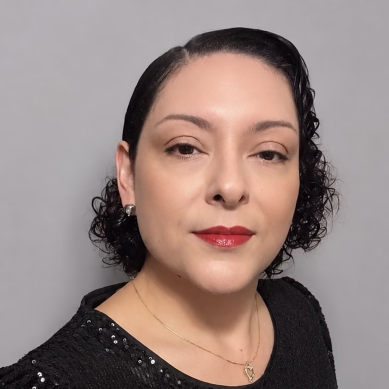 Portrait of a woman with short curly dark hair, red lipstick, wearing a black sequined top and a delicate gold necklace against a gray backdrop.
