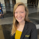 Smiling woman with long blond hair wearing a black blazer and yellow top in a modern lobby.