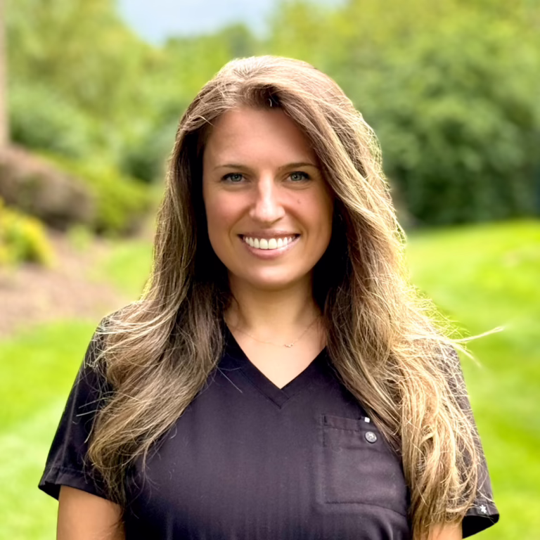 Smiling woman with long light-brown hair wearing black scrubs, standing outdoors in a green garden background.