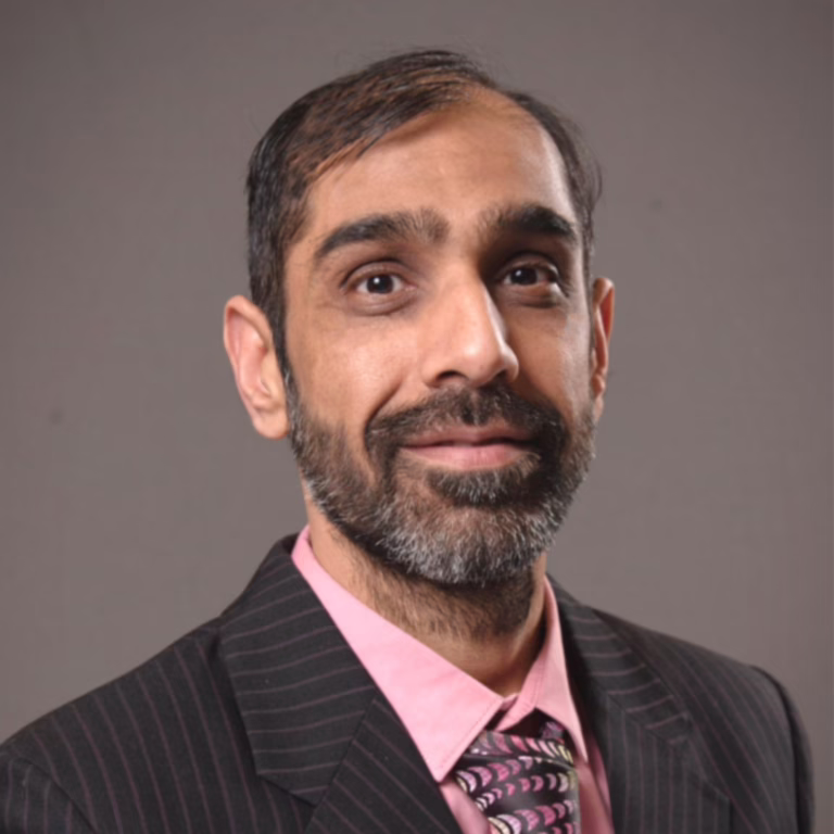Professional headshot of a man with dark hair and beard, wearing a pink shirt and pinstriped suit, looking at the camera with a gentle smile.