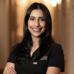 Portrait of a smiling woman with long black hair wearing a black dental uniform in a warmly lit hallway.