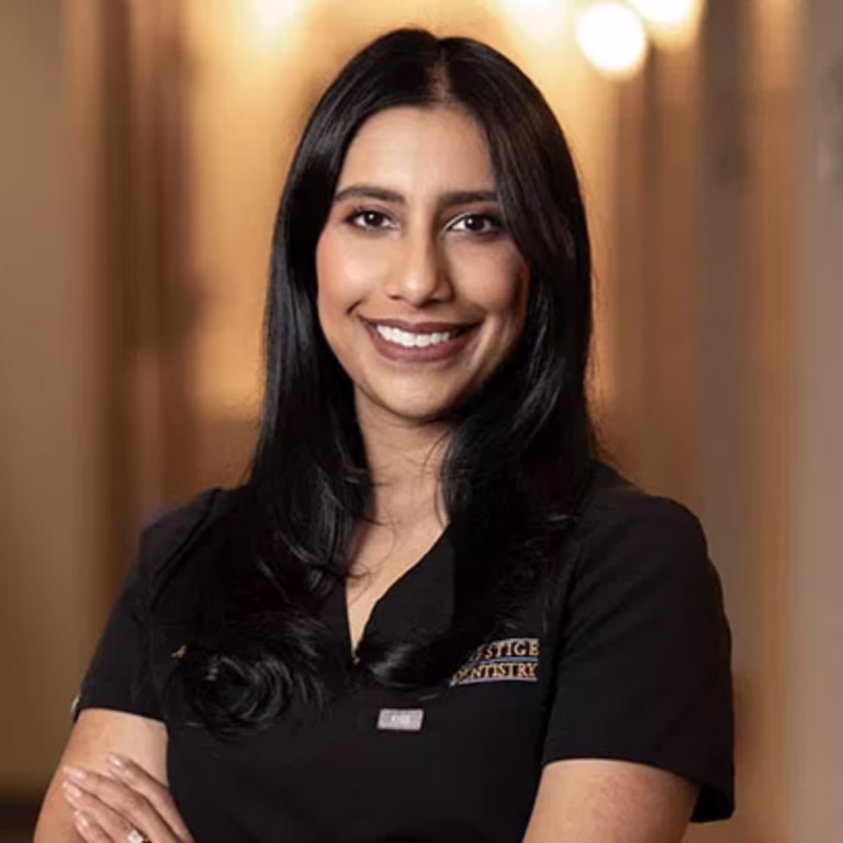 Portrait of a smiling woman with long black hair wearing a black dental uniform in a warmly lit hallway.