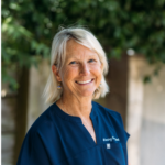 Smiling woman with short gray-blond hair in navy scrub top outdoors, greenery in the background.