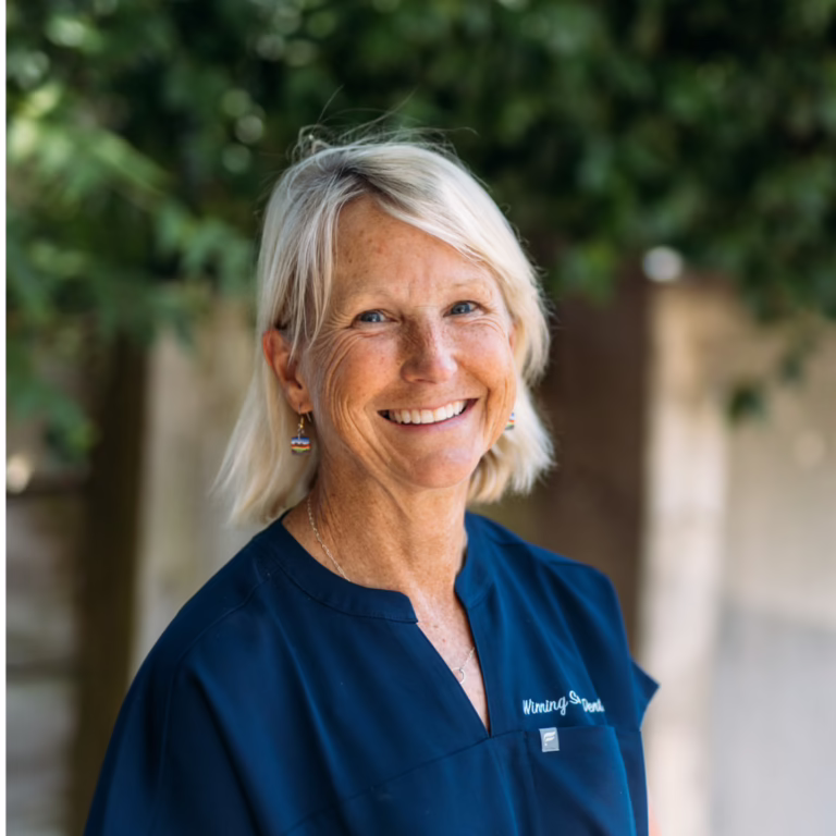 Smiling woman with short gray-blond hair in navy scrub top outdoors, greenery in the background.