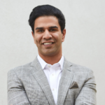 Smiling man in a light gray check blazer and white shirt, arms crossed, standing against a neutral wall with a pocket square showing.