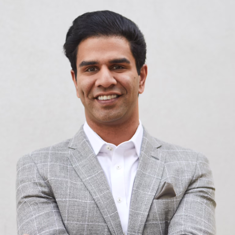 Smiling man in a light gray check blazer and white shirt, arms crossed, standing against a neutral wall with a pocket square showing.