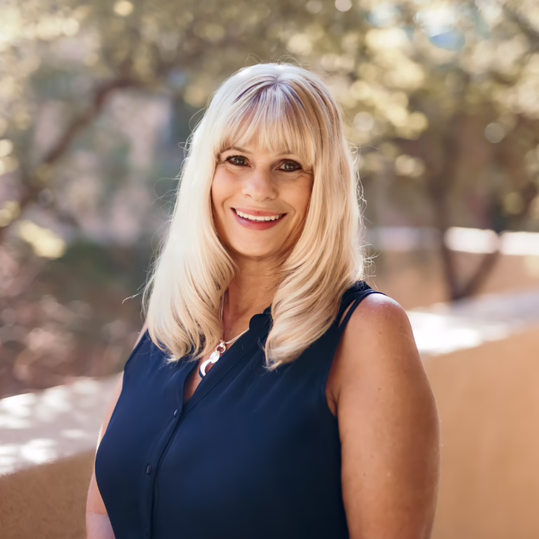 Smiling woman with long blonde hair in a navy sleeveless blouse outdoors, sunlit background.