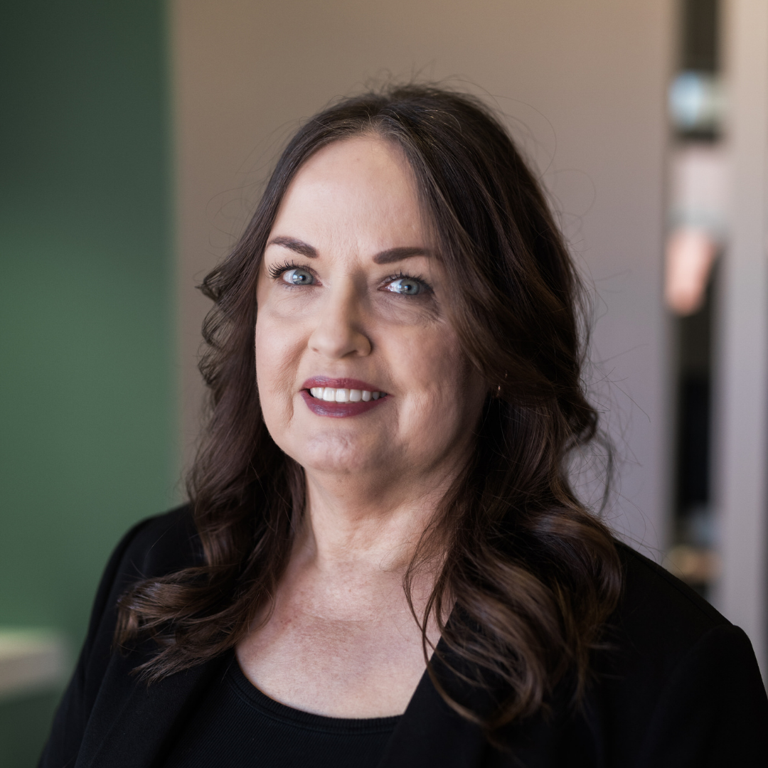 Portrait of a smiling woman with blue eyes and wavy brown hair, wearing a black top in an office setting.
