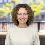 Smiling woman with curly hair in a light sweater, outdoors with a blurred background of trees and buildings.