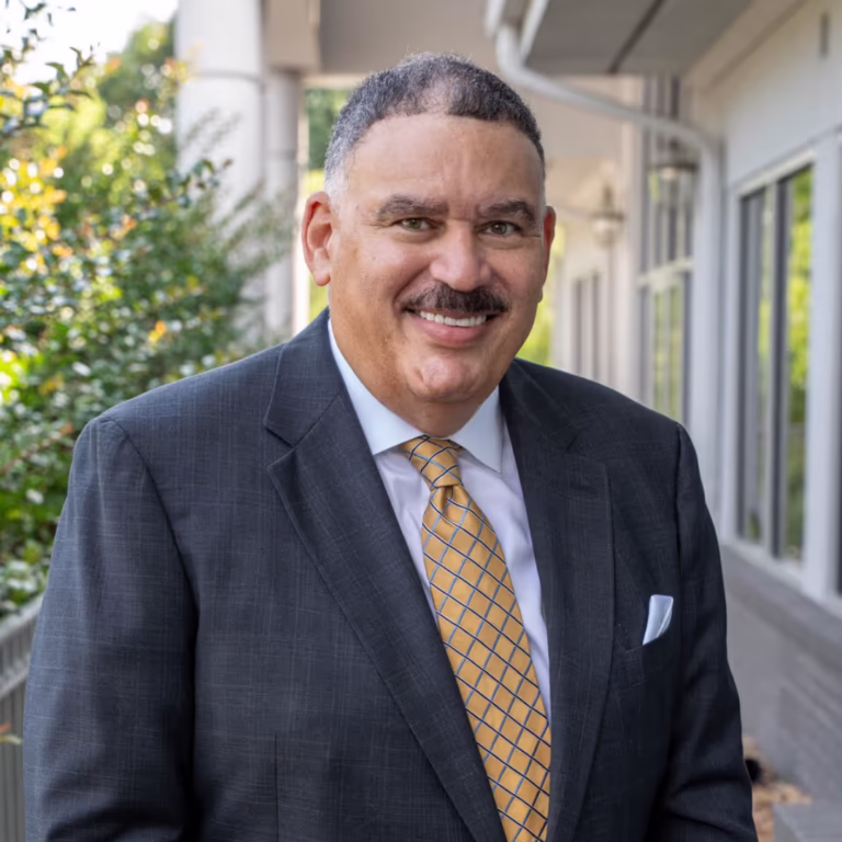 Smiling man in a dark suit and yellow tie stands outdoors near a building, posing for a professional headshot.