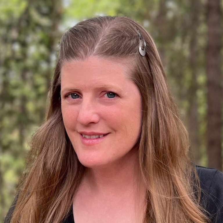 Smiling woman with long light brown hair and a hair clip, outdoors in a wooded area.