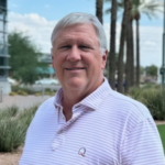 Smiling older man with white hair in a light striped polo shirt, outdoors with palm trees in the background.