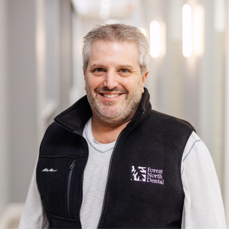 Smiling middle-aged man in a black vest with Forest North Dental logo, standing in a bright hallway.