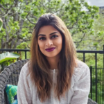 Portrait of a woman with long brown hair and purple lipstick, wearing a white textured top, sitting on a wicker chair on a balcony with green trees in the background
