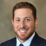 Smiling man in a dark blazer, light blue shirt, and blue tie—professional headshot.