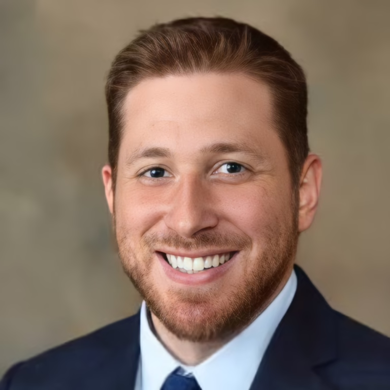 Smiling man in a dark blazer, light blue shirt, and blue tie—professional headshot.