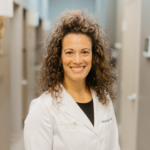 Smiling female dentist in a white coat standing in a clinic corridor.
