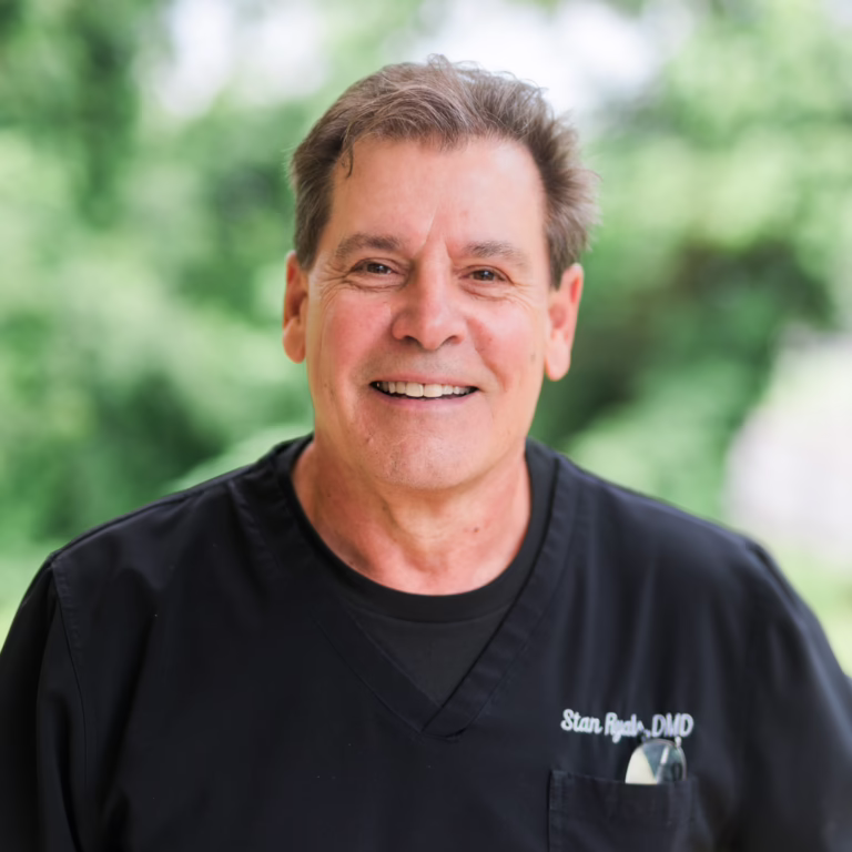 Smiling middle-aged man in black medical scrubs outdoors, name badge reads 'Stan R...' with glasses in a pocket on his chest pocket.