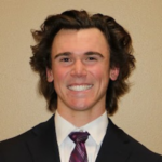 Young man with wavy brown hair in a navy suit and white shirt, smiles at the camera.